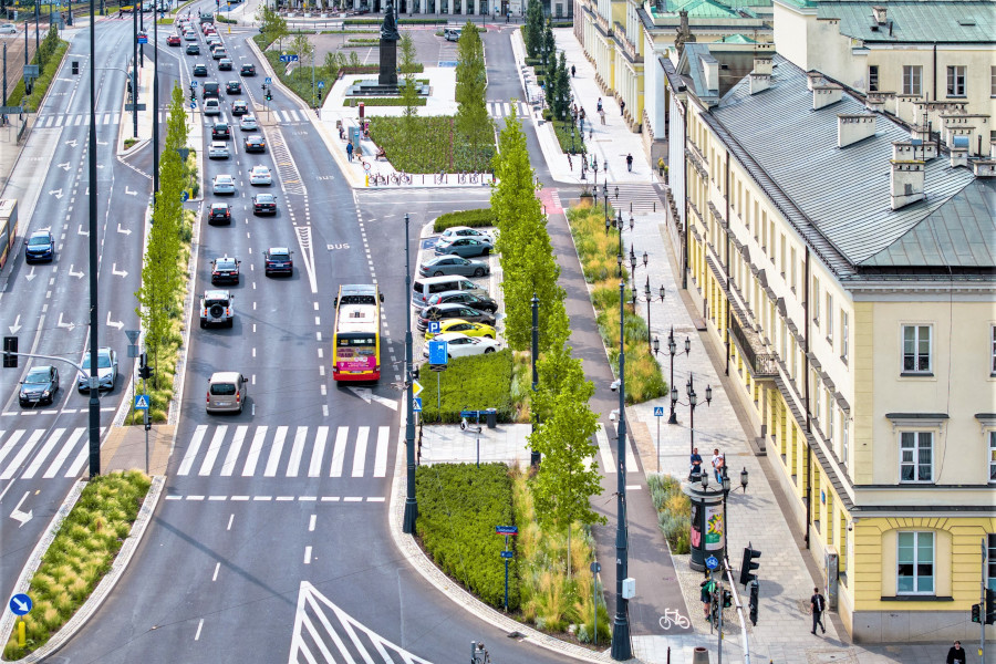 Ruchliwa ulica z samochodami i autobusem, obok zieleń oraz piesi spacerujący po chodniku w miejskim otoczeniu.