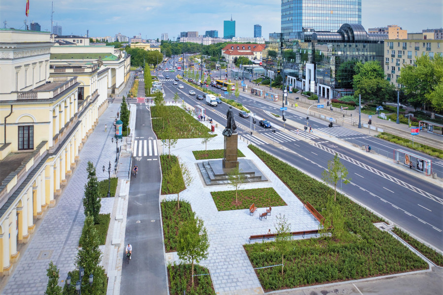 Zielony skwer z pomnikiem w centrum, otoczony nowoczesnymi budynkami i ulicami z ruchem pojazdów.