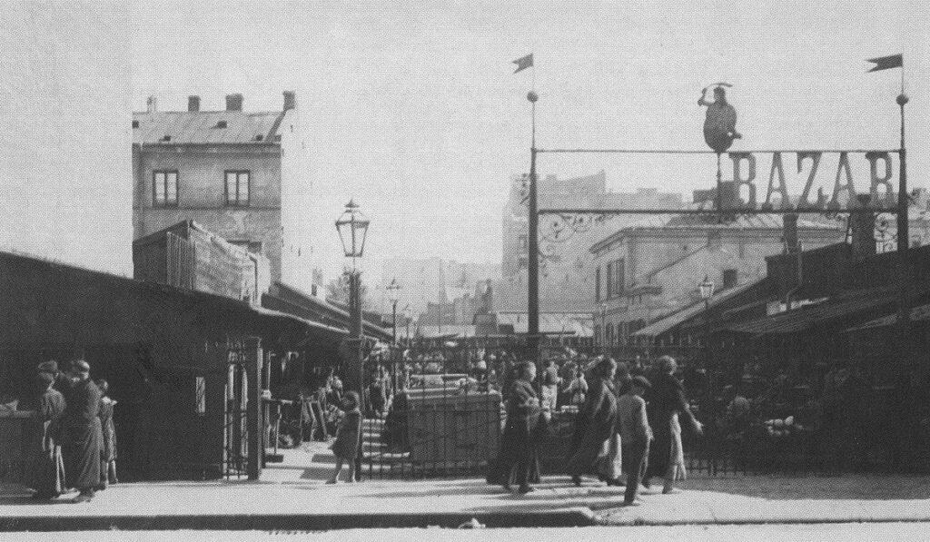 Tłum ludzi przechadza się pomiędzy straganami na bazarze, w tle widoczny szyld "BAZAR" oraz zabudowania.