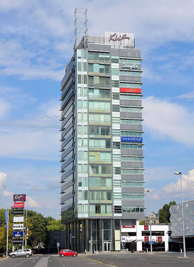 Nowoczesny biurowiec z przeszkloną fasadą, z logo "Klip" na dachu, otoczony parkingiem i sklepami.