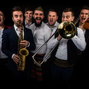 Grupa mężczyzn w tradycyjnych strojach i garniturach, grająca na instrumentach muzycznych, w tym trąbce i saksofonie, w ciemnym otoczeniu.