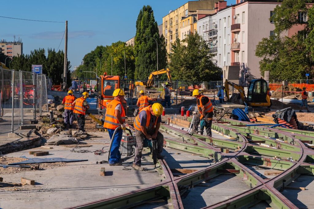 Grupa robotników w kamizelkach odblaskowych pracuje nad torami tramwajowymi w miejskim otoczeniu.