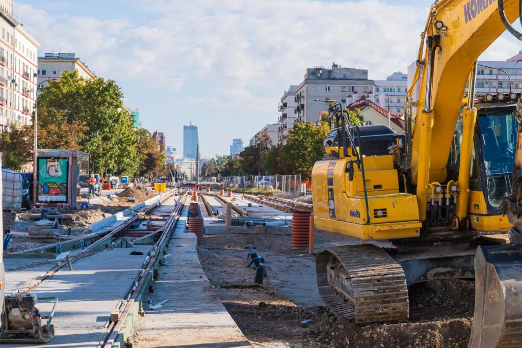 Żółty koparka pracująca na budowie torów tramwajowych w miejskim otoczeniu, w tle nowoczesne budynki.