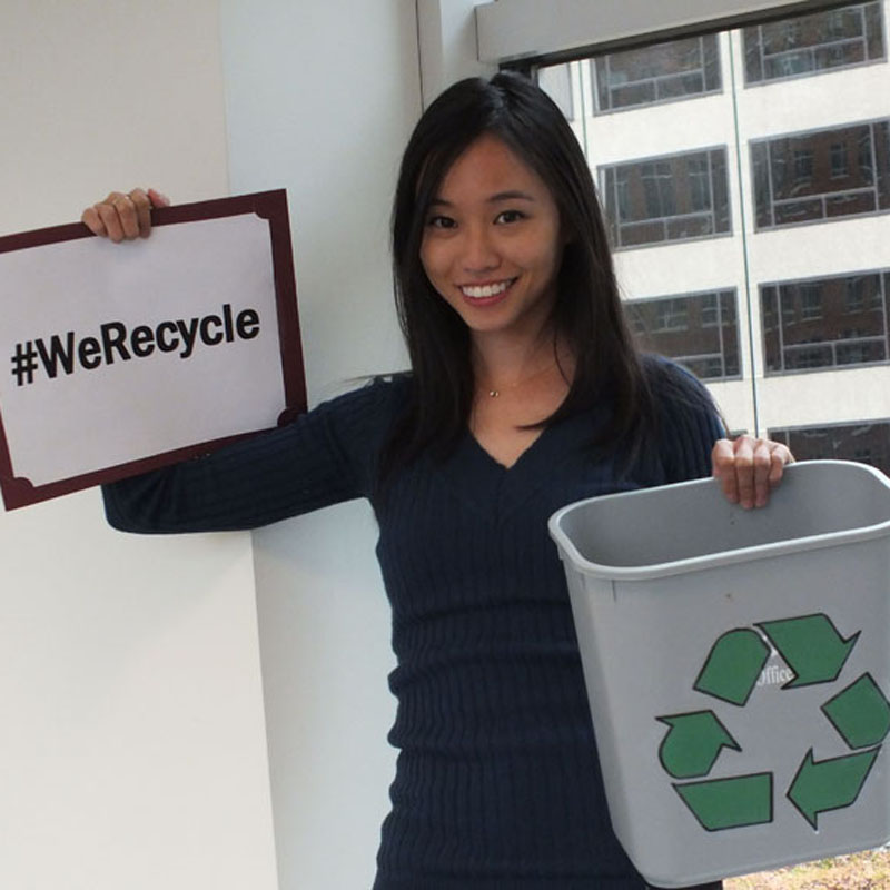 Kobieta w niebieskiej bluzie trzyma znak z napisem "#WeRecycle" oraz szary pojemnik do recyklingu w biurowym otoczeniu.