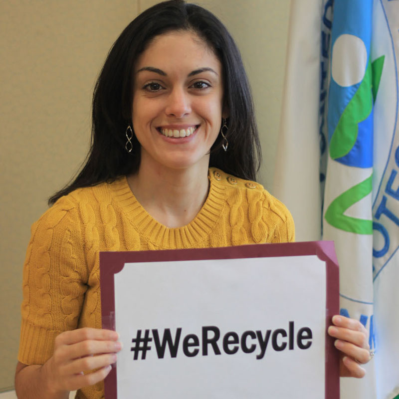 Kobieta w żółtym swetrze trzyma tabliczkę z napisem "#WeRecycle" w biurowym otoczeniu.