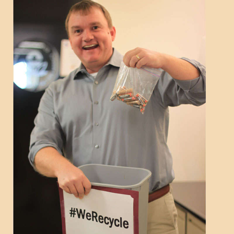 Mężczyzna w koszuli trzyma woreczek z bateriami, przygotowując się do ich wrzucenia do pojemnika z napisem "#WeRecycle".