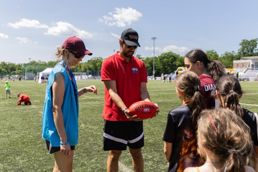 Mężczyzna w czerwonej koszulce trzyma piłkę, rozmawiając z grupą dzieci na boisku sportowym.