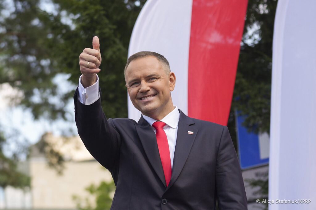 Mężczyzna w garniturze z uśmiechem podnosi kciuk w górę na tle flagi.