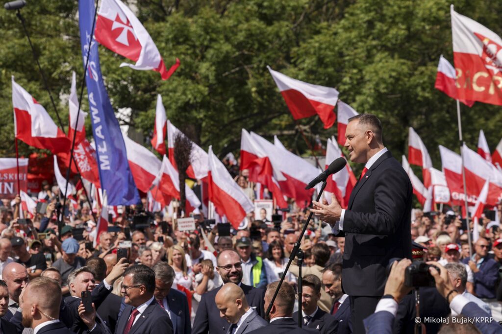 Mężczyzna w garniturze przemawia do tłumu, w tle powiewają polskie flagi.