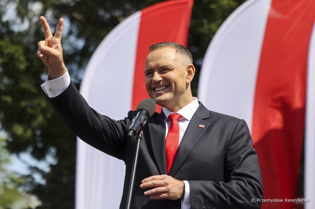 Uśmiechnięty mężczyzna w garniturze z mikrofonem na mównicy, pokazujący znak pokoju, w tle flagi narodowe.