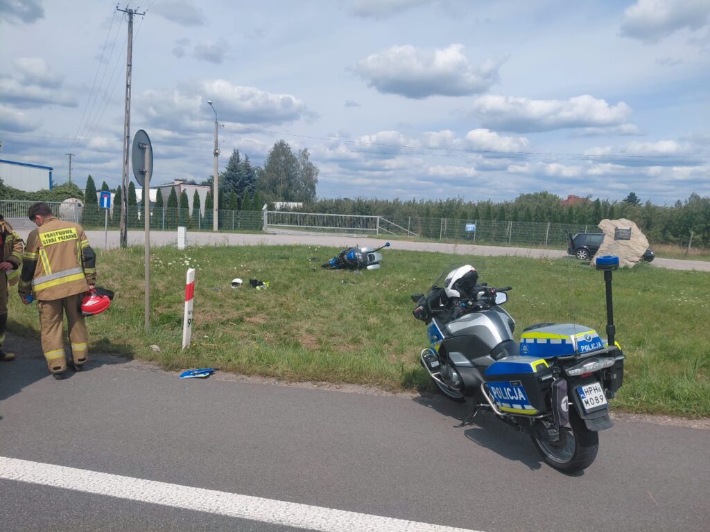 Dwaj strażacy w mundurach omawiają sytuację obok uszkodzonego motocykla na poboczu drogi.