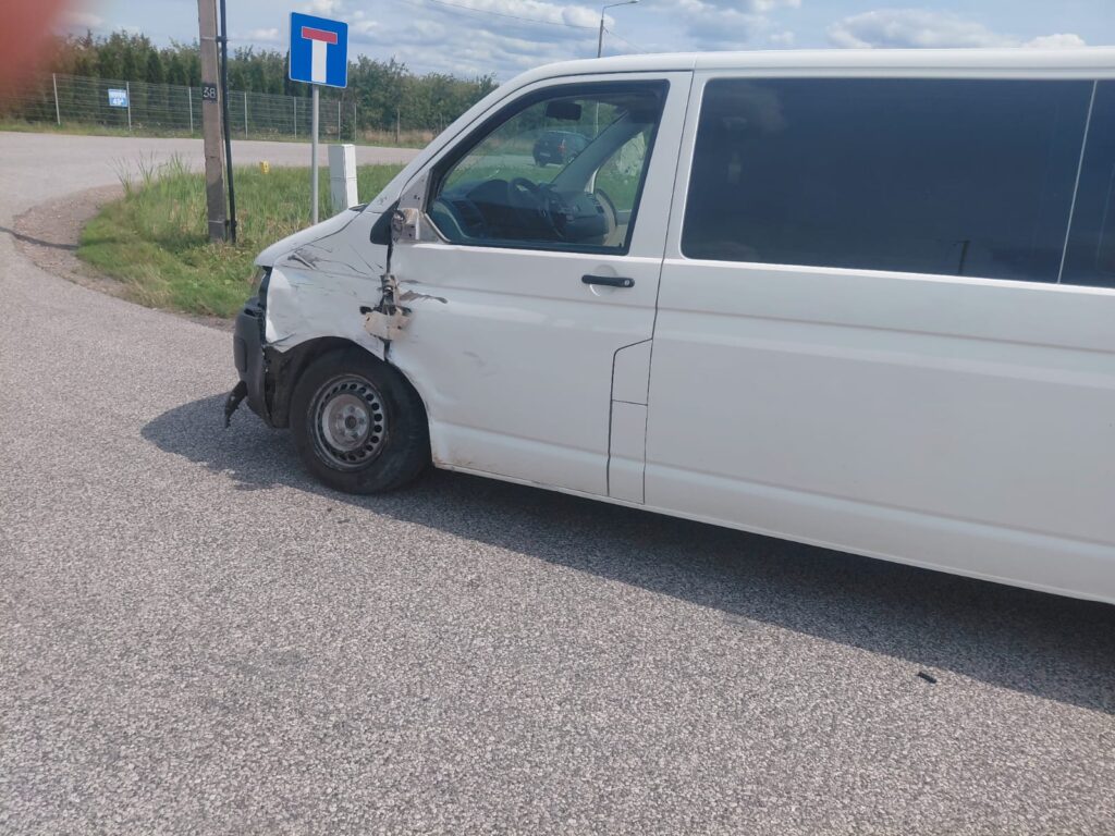 Biały van z uszkodzoną przednią częścią stoi na zakręcie drogi, obok znaku drogowym.
