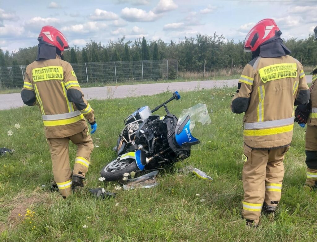 Strażacy w hełmach oceniają przewrócony motocykl na poboczu drogi, w tle zieleń i ogrodzenie.