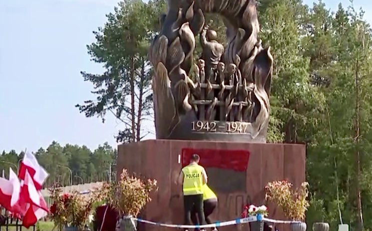 Mężczyzna w kamizelce policyjnej odsłania pomnik z postaciami, otoczony flagami i kwiatami w lesistym terenie.