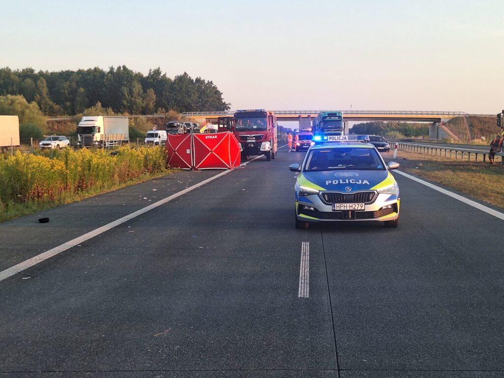 Policyjny radiowóz z włączonymi sygnałami świetlnymi stoi na drodze, obok widoczna czerwona osłona, a w tle pojazdy służb ratunkowych.