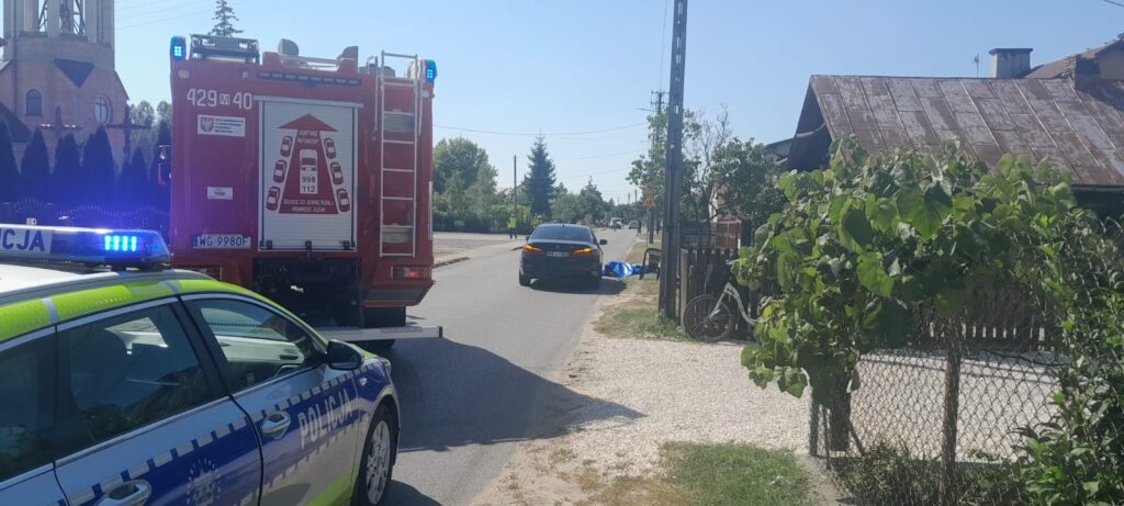 Na drodze stoi radiowóz policyjny i wóz strażacki, obok leży niebieski koc. W tle widoczna wieża kościoła.