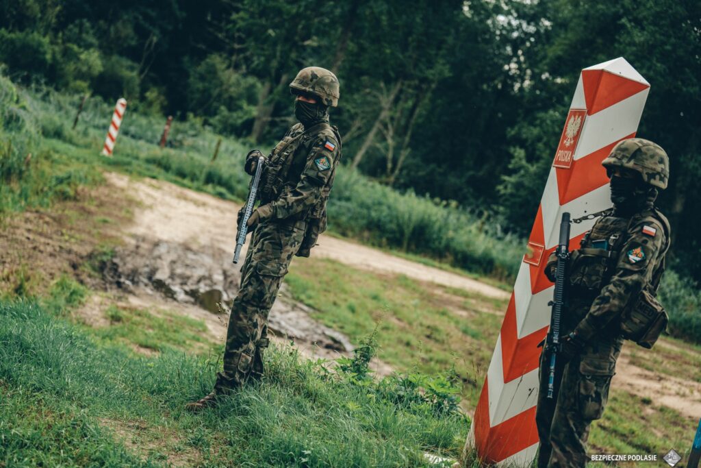 Dwaj żołnierze w mundurach kamuflażowych stoją na straży przy oznakowanej słupie granicznym w zalesionym terenie.