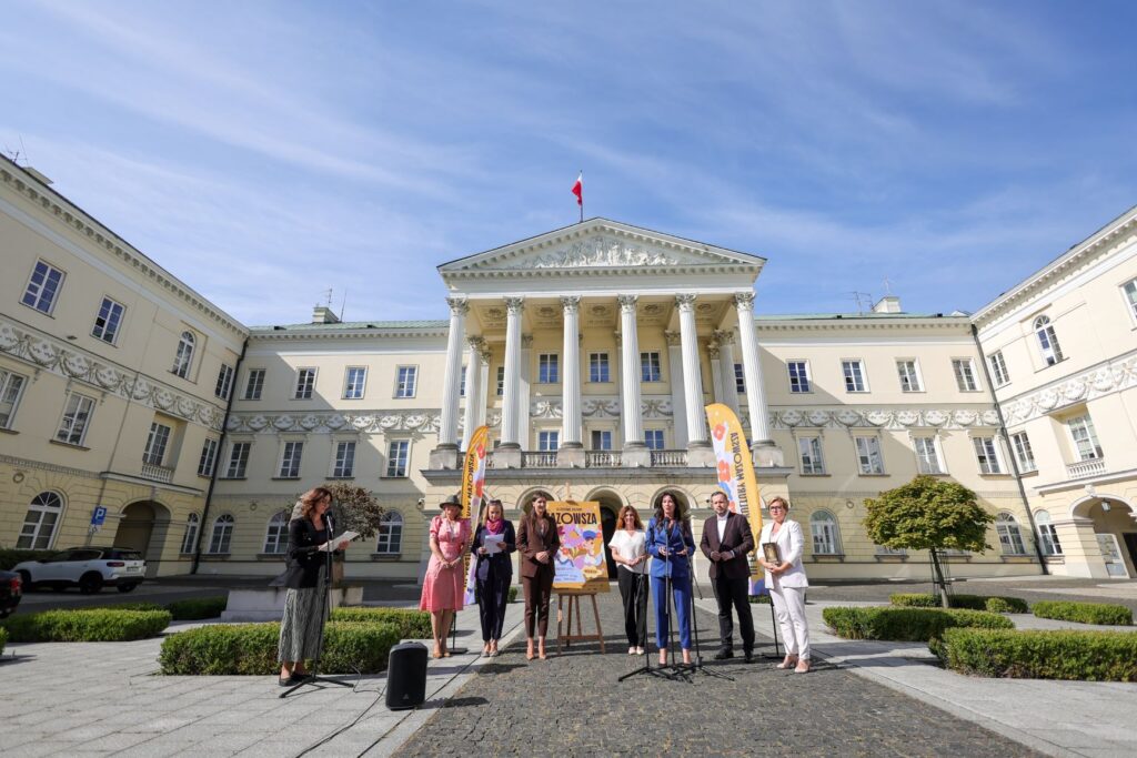 Kobieta w garniturze przemawia do mikrofonu przed budynkiem z kolumnami, a obok niej stoją inne osoby z dokumentami.