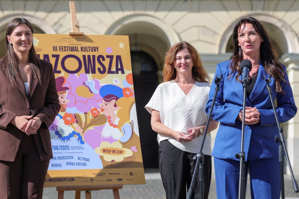 Trzy kobiety stoją przy mównicy, obok plakatu III Festiwalu Kultury Mazowsza, w słoneczny dzień na tle budynku.