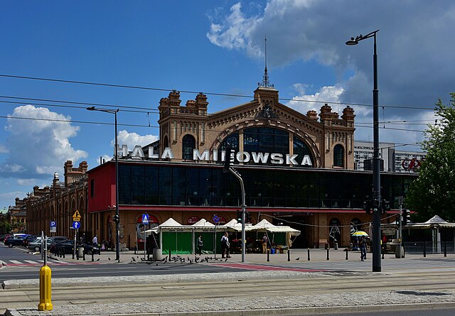 Hala Mirowska z charakterystyczną, zabytkową architekturą i przeszkloną fasadą, otoczona ulicami i zielenią.