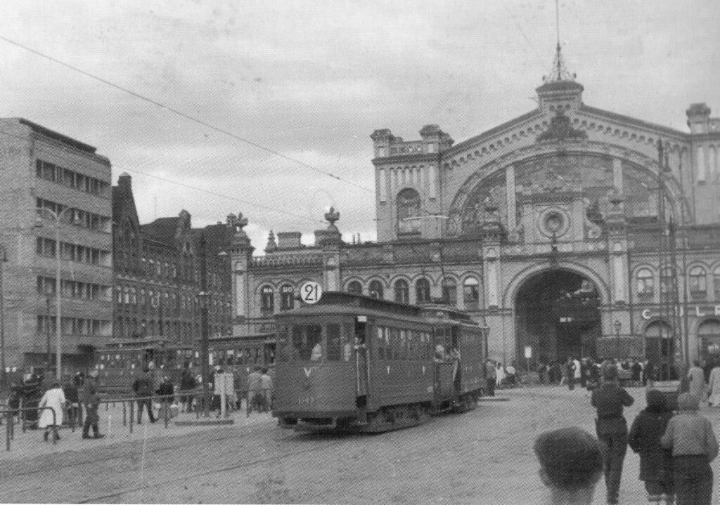 Tramwaj z numerem 21 przejeżdża przed zabytkowym budynkiem, w tle widoczni spacerujący ludzie.