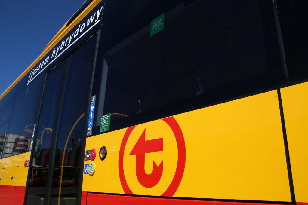 Hybrydowy autobus z logo przewoźnika, stojący na przystanku w słoneczny dzień.