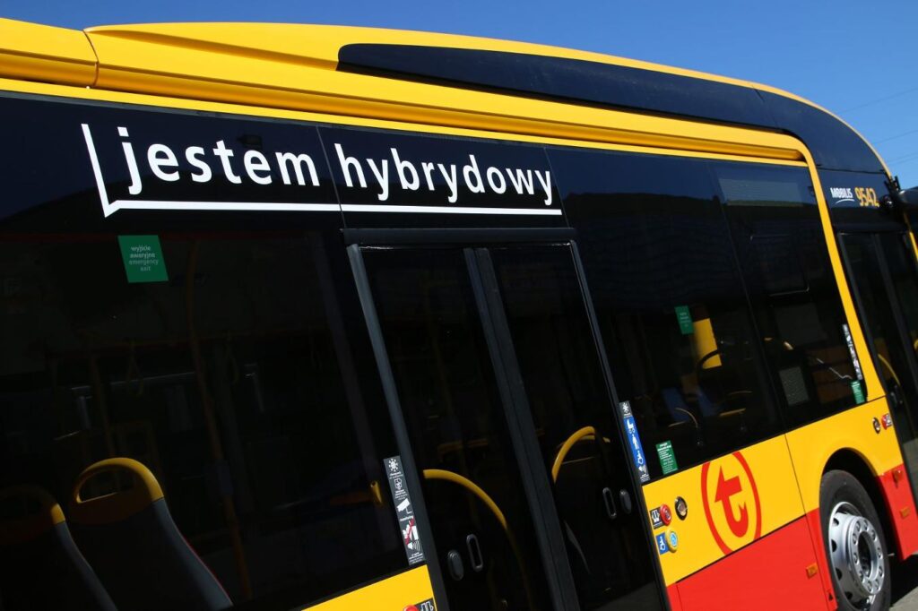 Hybrydowy autobus z napisem „jestem hybrydowy” na bocznej szybie, zaparkowany w słonecznym otoczeniu.