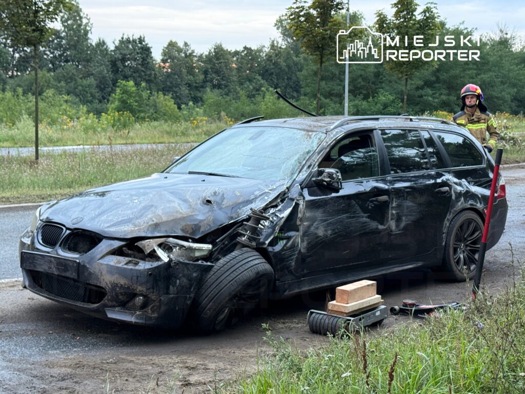 Uszkodzony czarny samochód osobowy stoi na poboczu drogi, obok niego stoi strażak w hełmie.