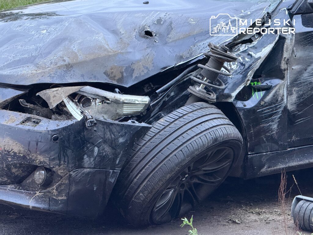 Uszkodzony czarny samochód osobowy leży na boku, z wygiętą karoserią i zniszczoną przednią lampą.