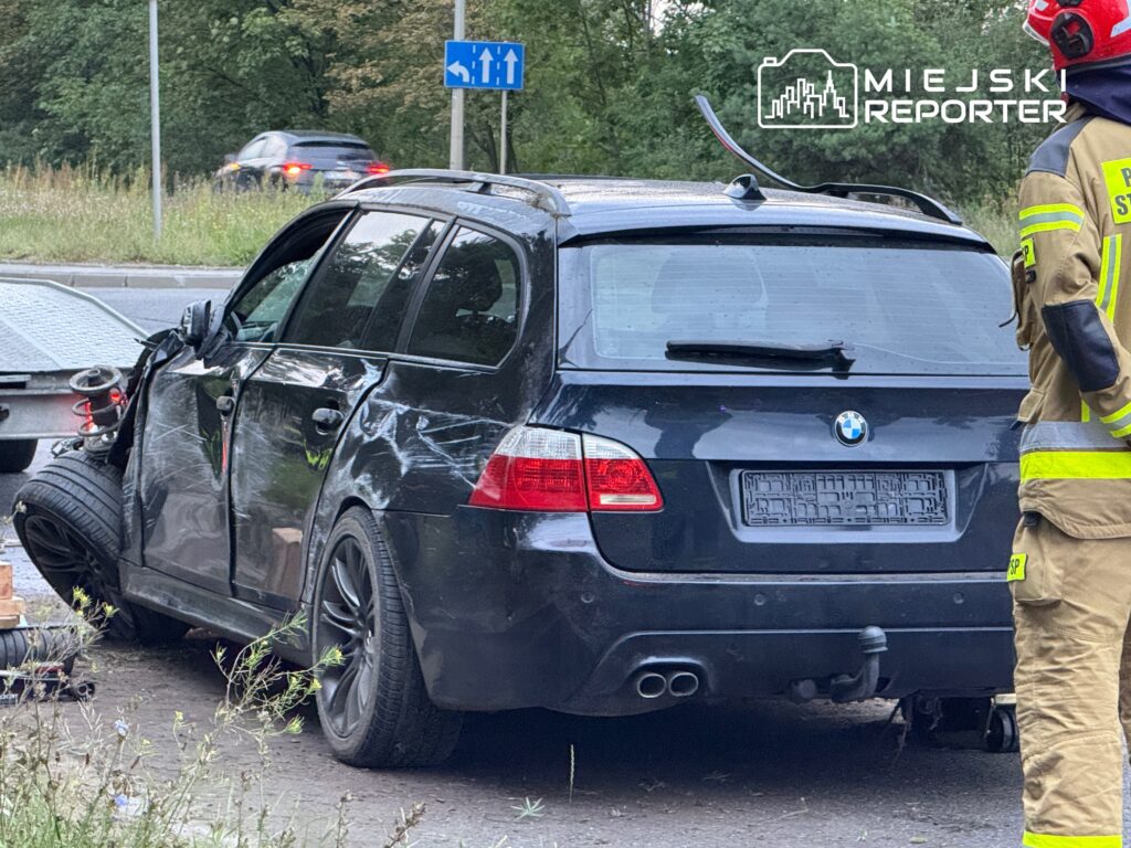 Czarny samochód osobowy z uszkodzeniami bocznymi stoi na poboczu drogi, obok strażak w stroju ochronnym.