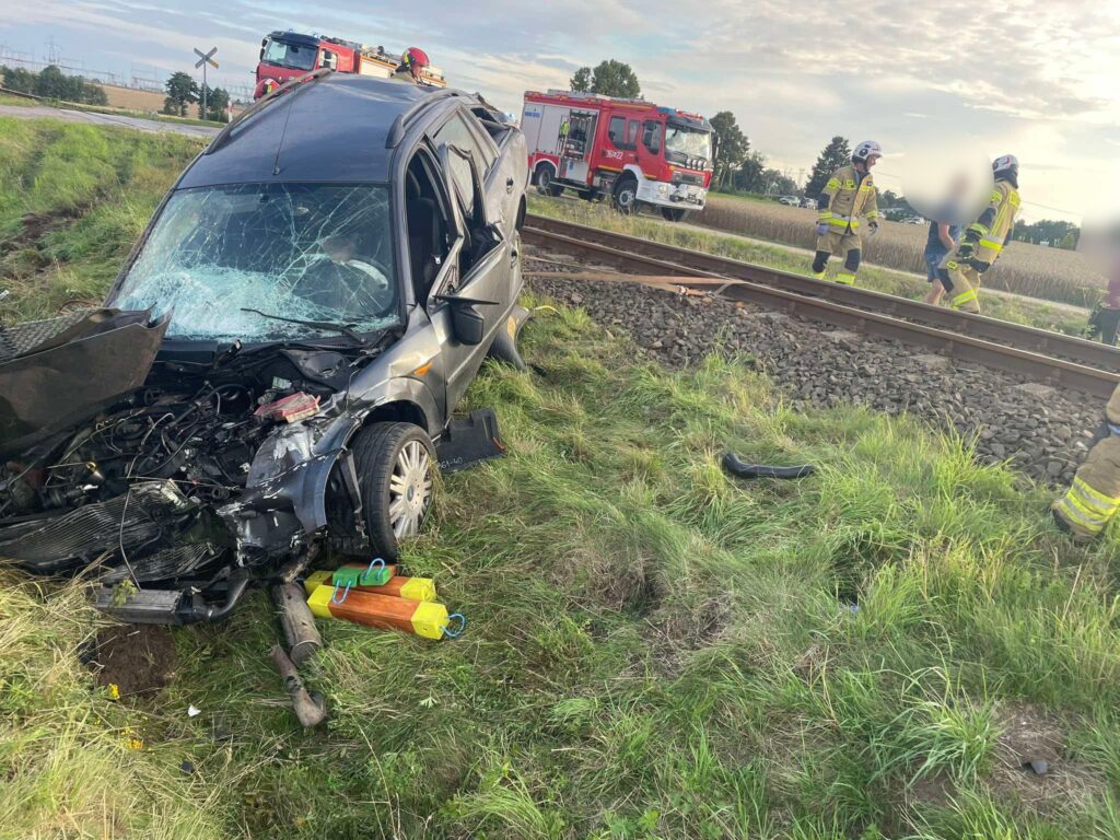 Uszkodzony samochód leży na poboczu drogi obok torów kolejowych, a strażacy udzielają pomocy osobie poszkodowanej.