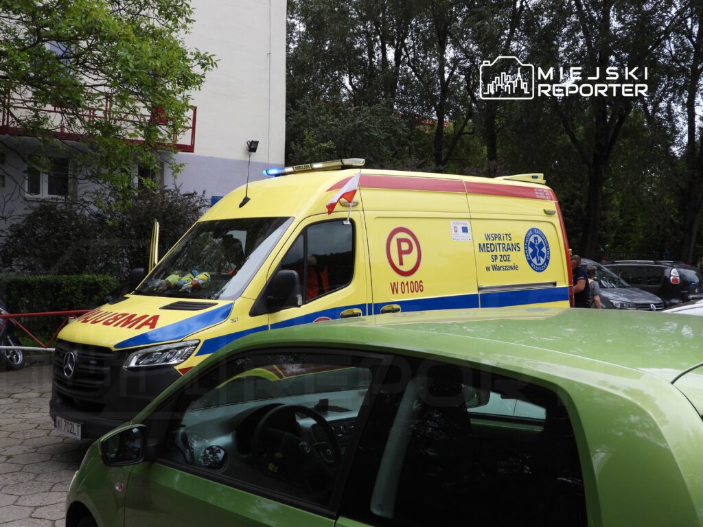 Karetka pogotowia z włączonymi sygnałami świetlnymi stoi na parkingu otoczonym drzewami.