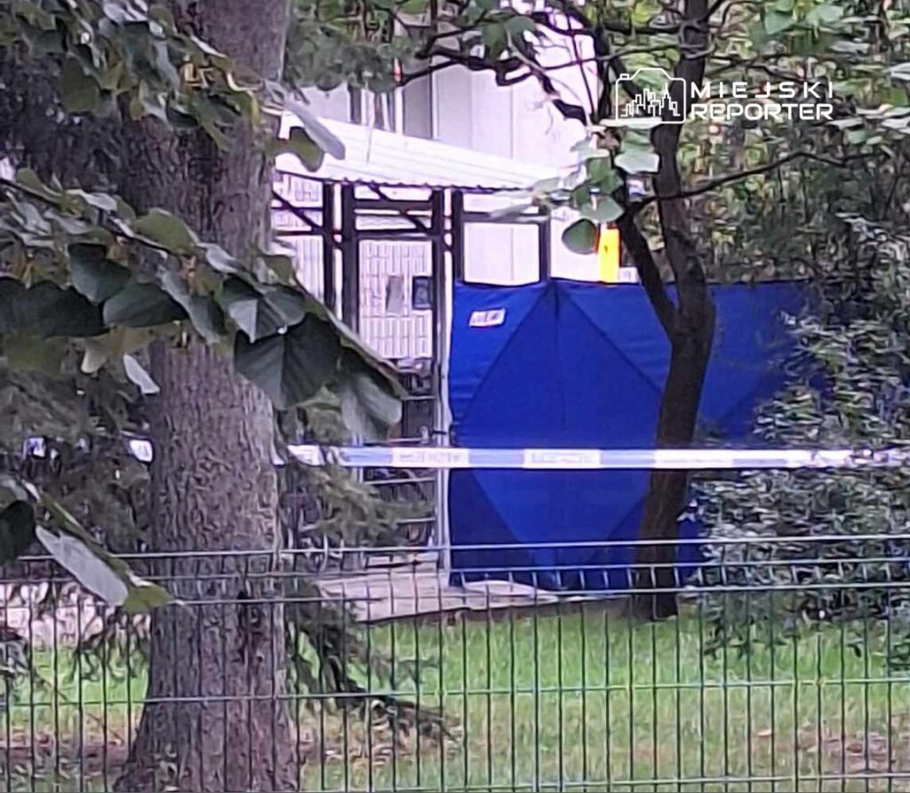 Nieoznakowane miejsce z niebieską osłoną, otoczone zielenią, zabezpieczone taśmą policyjną.