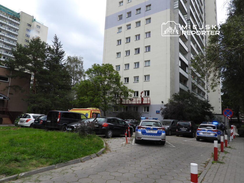 Na parkingu przed budynkiem stoją dwa radiowozy z włączonymi sygnałami świetlnymi, a obok nich zaparkowana karetka pogotowia.