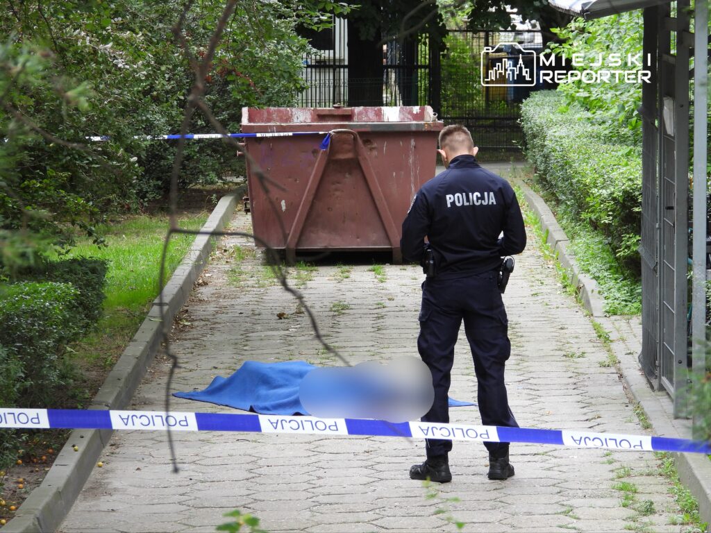 Policjant w mundurze bada miejsce zdarzenia przy kontenerze na śmieci, zabezpieczonym taśmą policyjną.