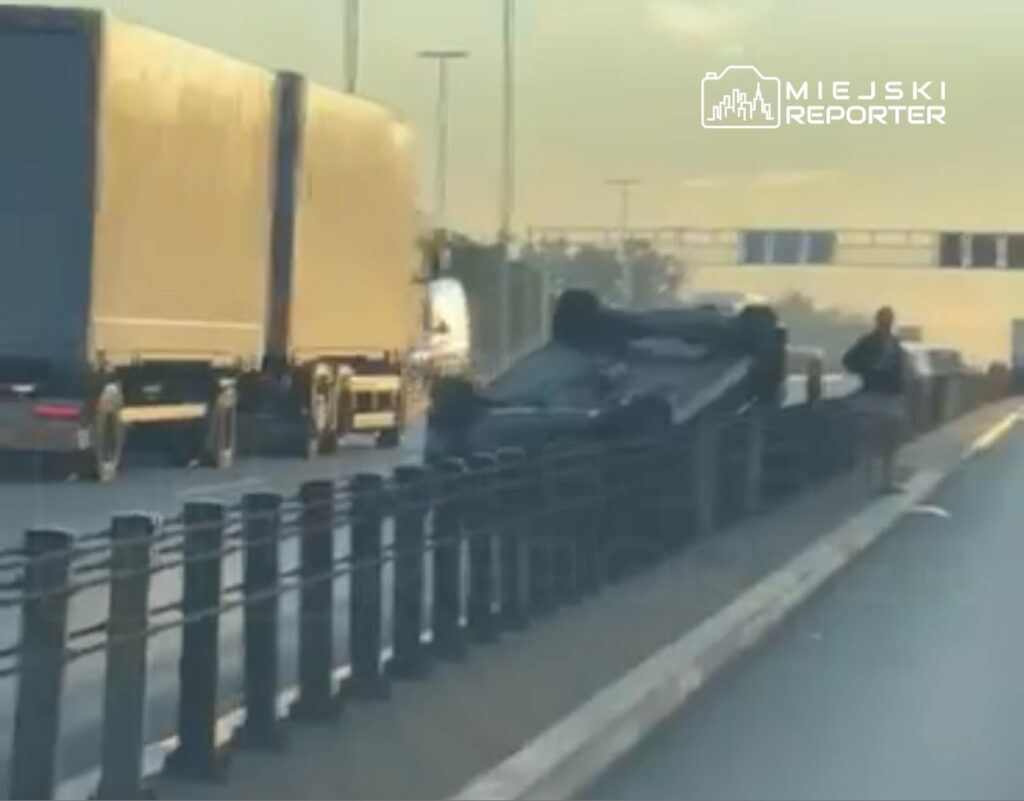 Na drodze leży przewrócone auto, obok stoi mężczyzna, a w tle przejeżdżają ciężarówki.