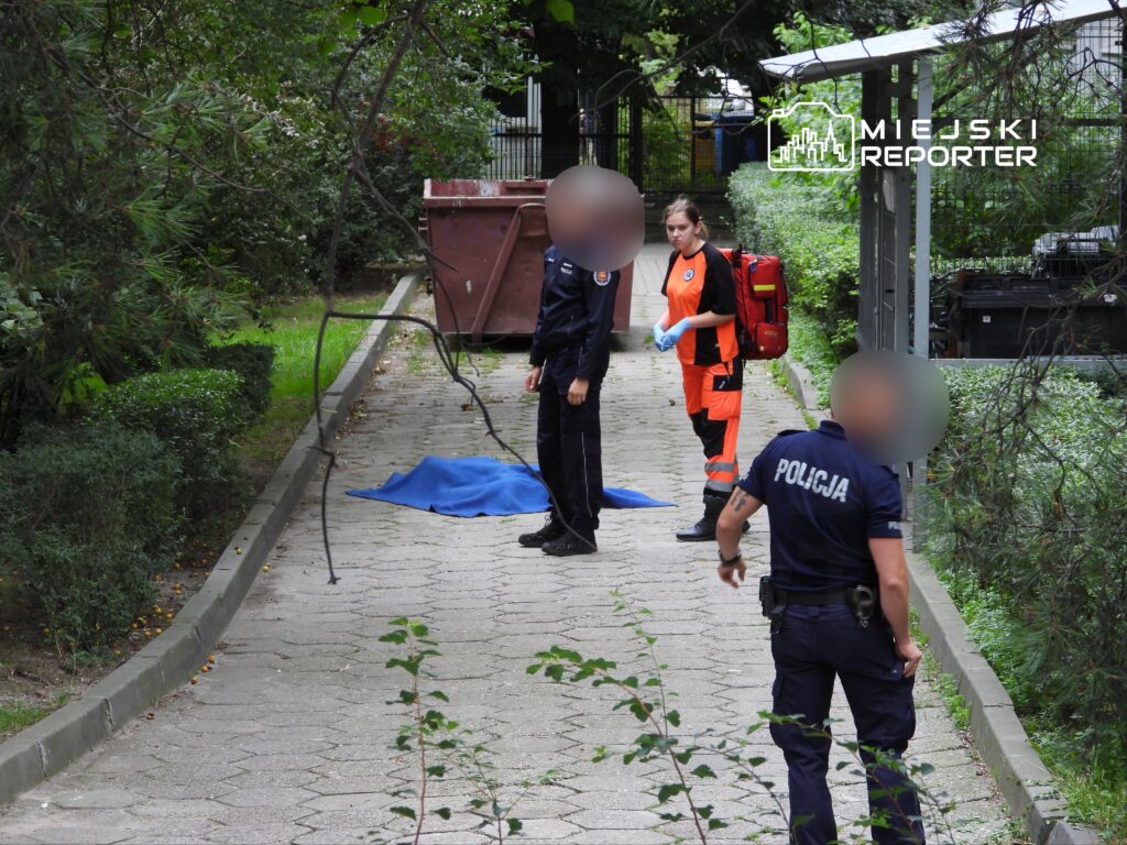 Ratowniczka w pomarańczowym stroju oraz policjanci na ścieżce w parku, obok niebieskiego koca.