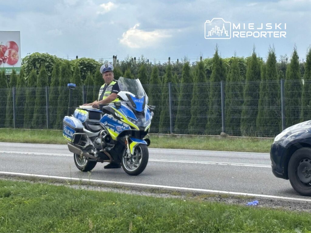 Policjant na motocyklu kontrolujący ruch drogowy przy asfaltowej drodze, w tle gęste krzewy i billboard z jabłkami.