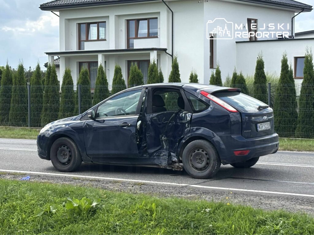 Uszkodzony niebieski samochód osobowy stoi na poboczu drogi, obok nowoczesnego budynku mieszkalnego.