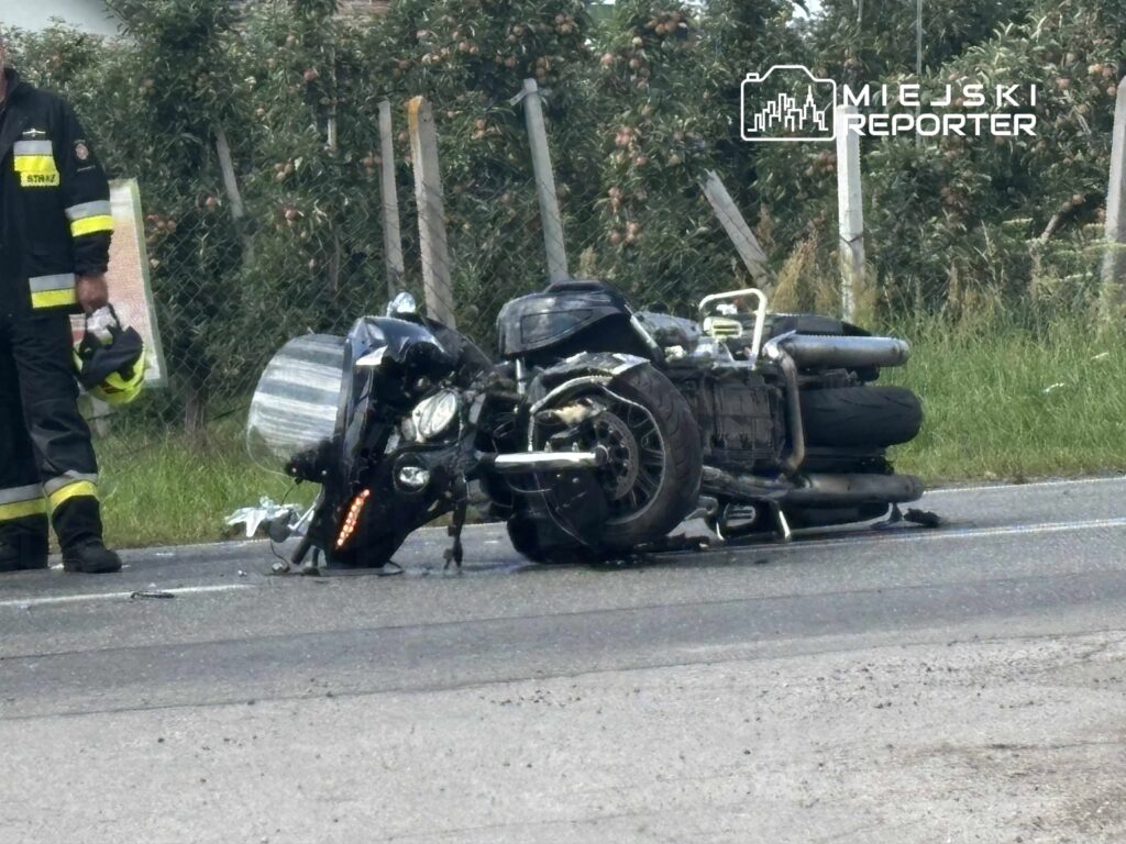 Strażak w mundurze stoi obok przewróconego motocykla na drodze, w tle widać zarośla.