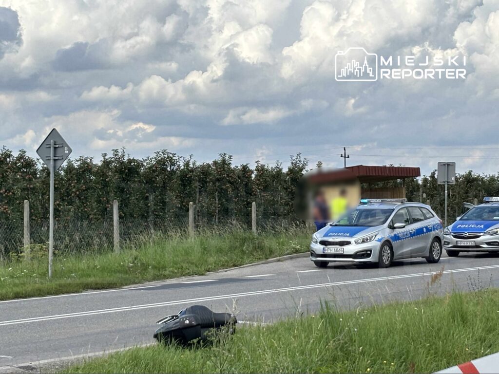 Policyjne samochody zaparkowane przy drodze, obok leżącego na jezdni motocykla, w tle gęsty sad.