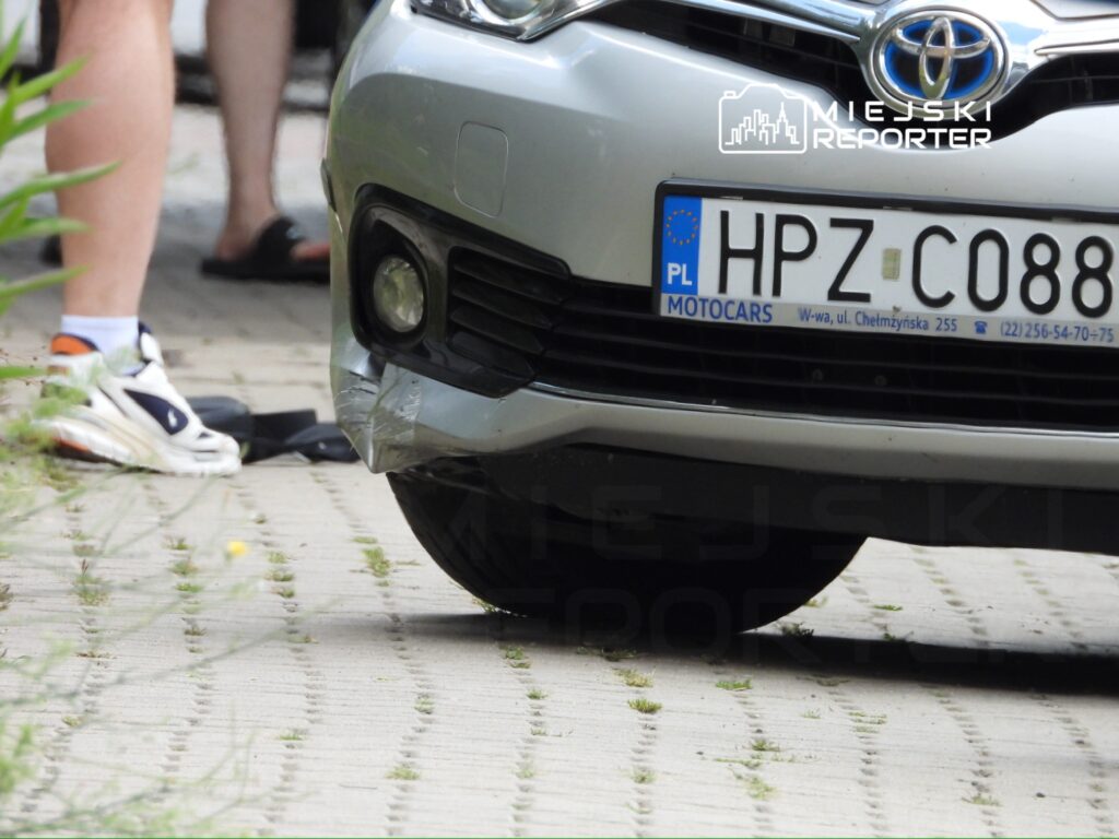 Samochód osobowy marki Toyota z uszkodzoną przednią częścią zaparkowany na chodniku w otoczeniu zieleni.