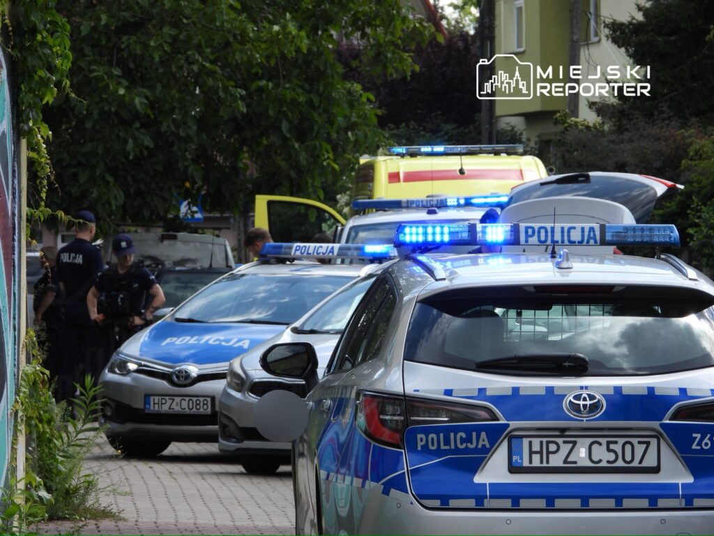 Grupa policjantów prowadzi interwencję w wąskiej uliczce, obok zaparkowanych radiowozów.