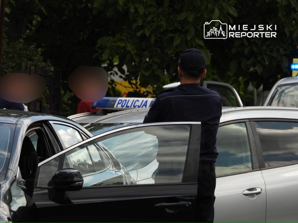 Policjant rozmawia z osobami stojącymi obok zaparkowanych samochodów w miejskim otoczeniu.