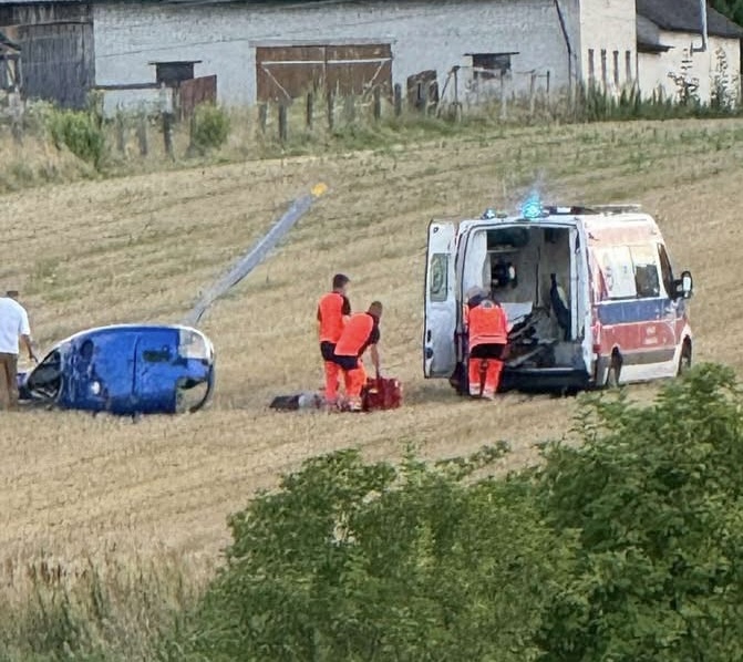 Ratownicy w pomarańczowych strojach udzielają pomocy przy przewróconym niebieskim pojeździe na polu.