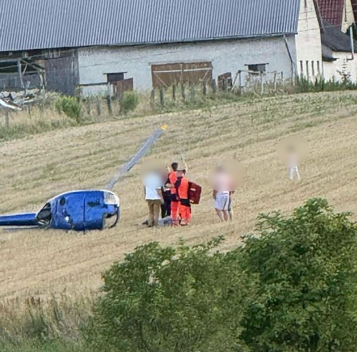 Zespół ratunkowy w pomarańczowych strojach udziela pomocy na polu obok przewróconego niebieskiego helikoptera.