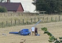 Na polu stoi przewrócony niebieski śmigłowiec, obok dwóch osób zajmujących się jego naprawą.