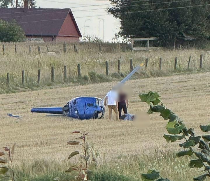 Na polu stoi przewrócony niebieski śmigłowiec, obok dwóch osób zajmujących się jego naprawą.