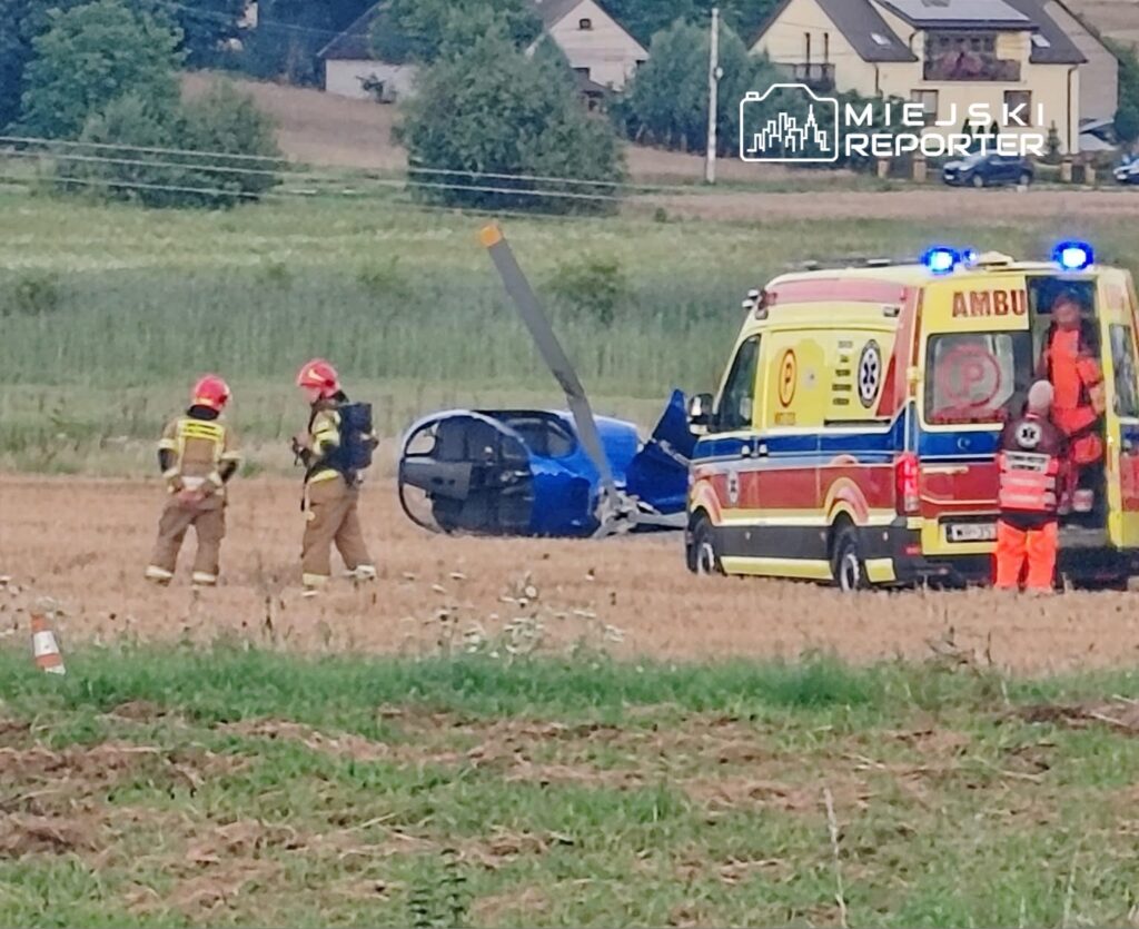 Strażacy w mundurach i kaskach rozmawiają obok przewróconego niebieskiego samochodu na polu, w tle karetka.