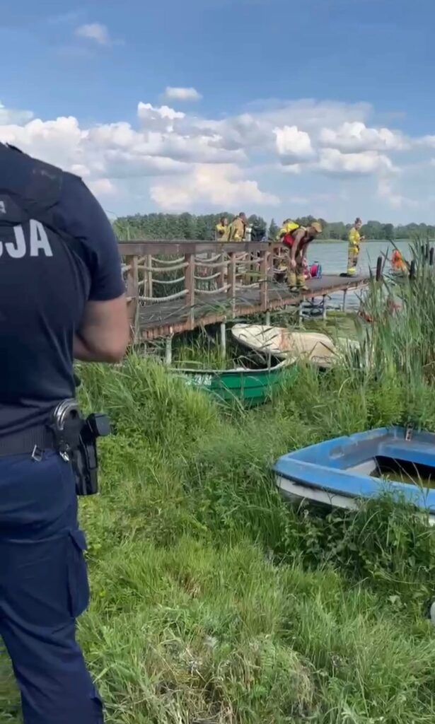 Policjant obserwujący akcję ratunkową na pomoście, gdzie strażacy przygotowują sprzęt nad wodą.